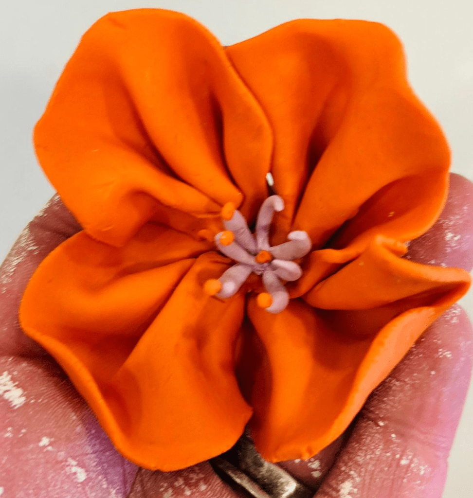 4. A close up of a hand holding a beautiful bright orange flower with lilac stamen, sculpted in modelling clay.