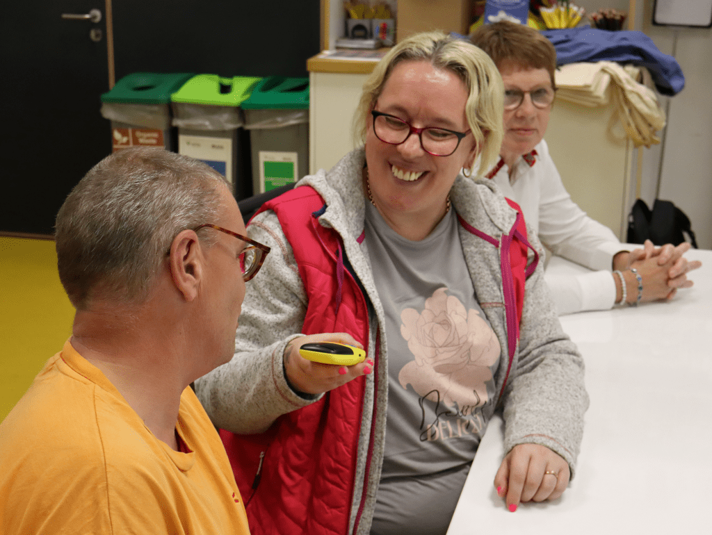 A blonde lady wearing pinky red glasses is smiling broadly. She is seated at a white table, holding a yellow audio recording device towards the face of a grey haired man as if interviewing him.
