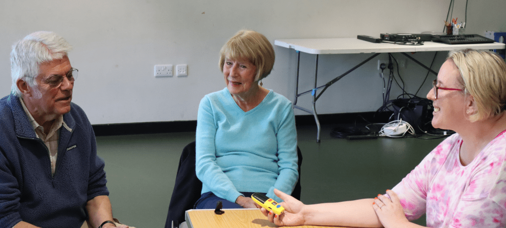 Three people are seated at a table in a  large studio with dark green floor and white walls. On the right, a blonde woman wearing a pink top and glasses smiles as she holds a yellow recording device out towards a grey haired man in a blue top and glasses. He is talking animatedly. In between the pair, an older lady with fair hair in a neat bob is listening intently and smiling.