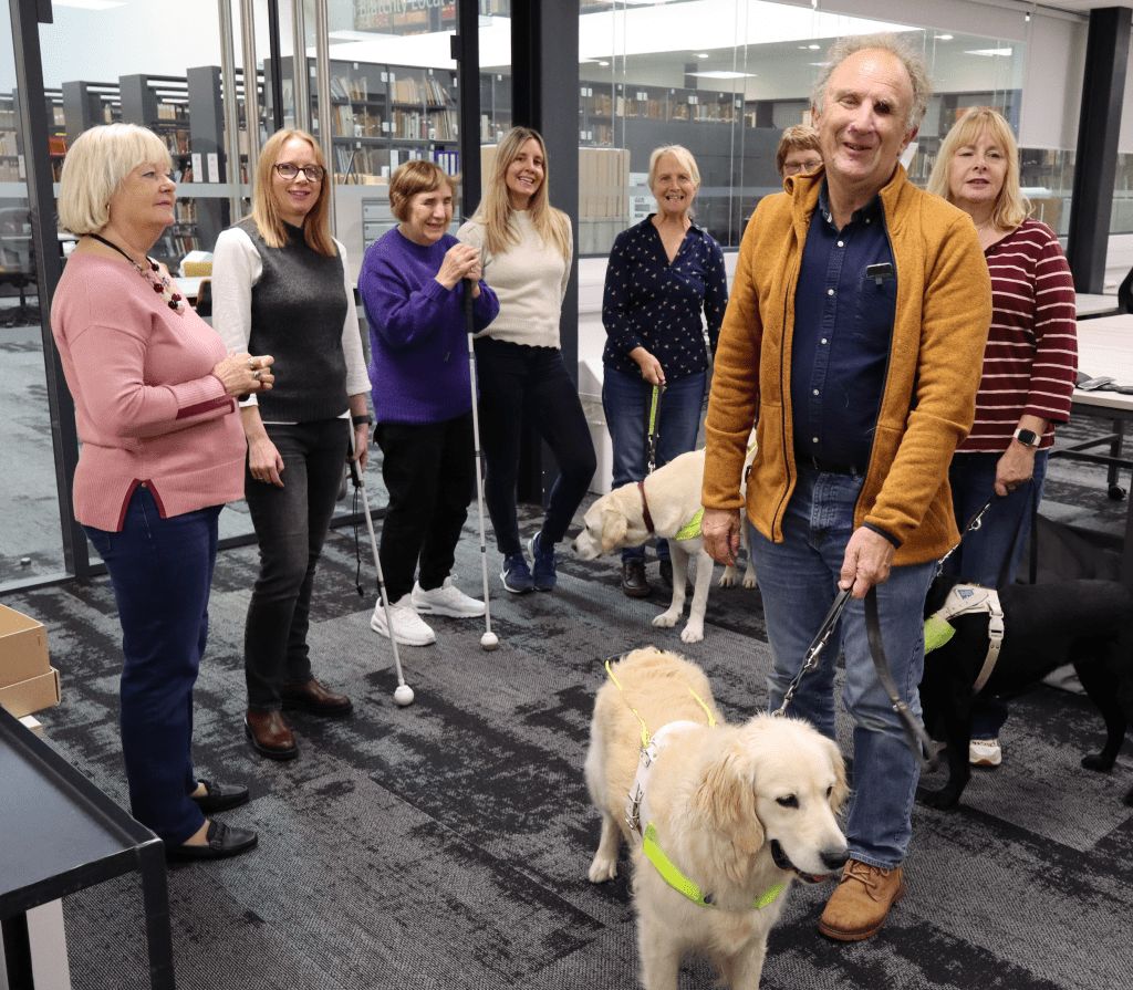 Eight people are standing in a carpeted office in front of a glass wall. Behind them through the glass wall a row of tall bookshelves are visible. Two of the people are holding white canes and three have guide dogs on leads. All are smiling.