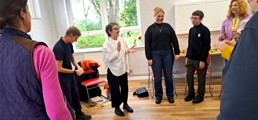 A woman with short brown-grey hair in a white shirt and black trousers stands in the centre of a group of a community space with white walls and a wooden floor. She is holding her hands in front of her, clapping. Around her is a circle of chairs, in front of each is a young person or adult who are watching and listening to her intently. Through a large window a garden scene can be seen behind them.