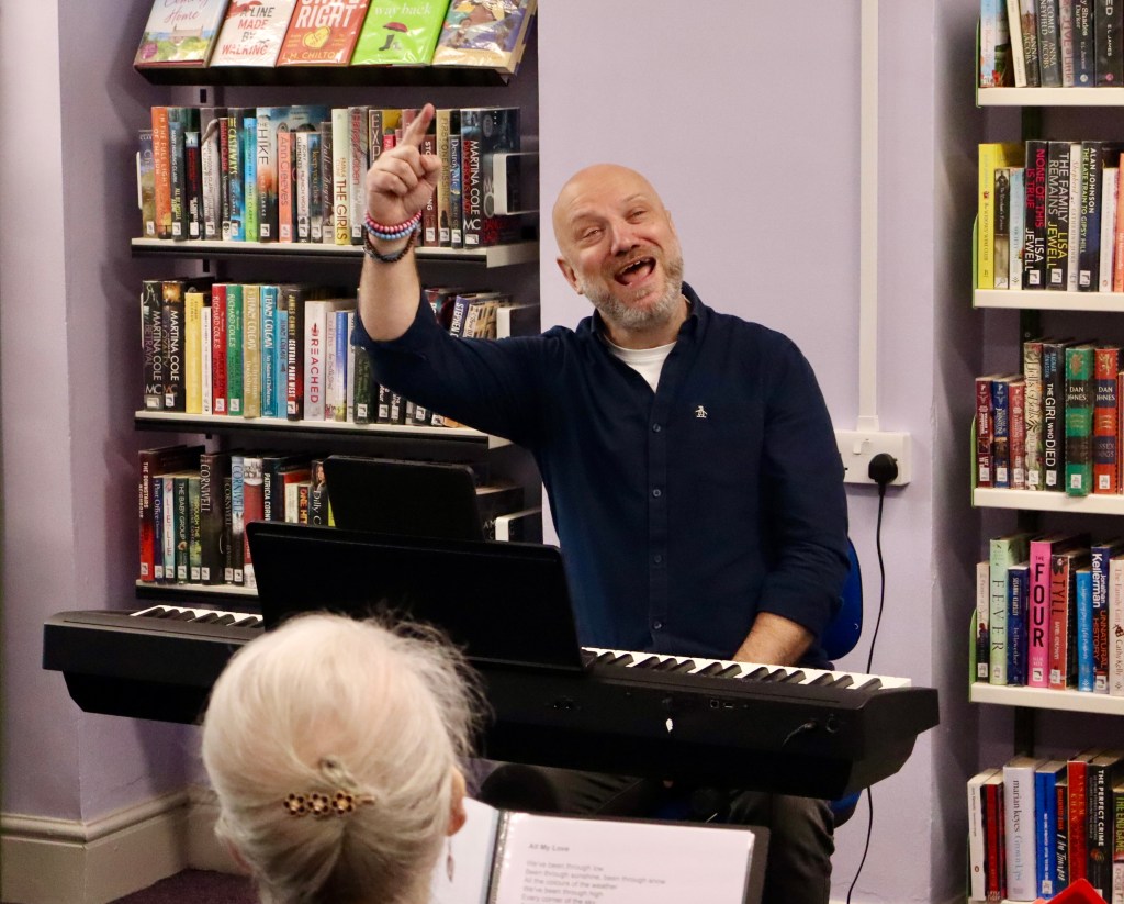 A man sits at a piano keyboard in a library. His hand is raised as he conducts a group of singers.